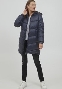 Marinblå pufferjacka med huva, dragkedjestängning, horisontellt quiltat mönster och justerbara snoddar, i kombination med svarta jeans och vita sneakers.