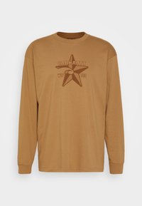 Pitkähiha-paita vaaleanruskeana, pehmeää puuvillaa, jossa on tähtigrafiikka ja "CARHARTT WIP 313" -teksti tummemmassa ruskeassa.