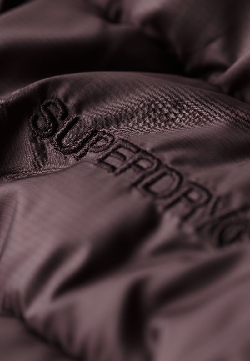 Ciemnofioletowa tkanina o miękkiej teksturze, z haftowanym logo "SUPERDRY" w dopasowanym kolorze. Widoczny jasny wzór siatki.