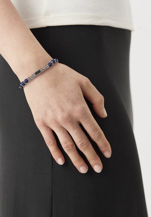 Poignet portant un bracelet en perles avec des perles bleues et argentées, main reposant sur un vêtement en tissu noir.