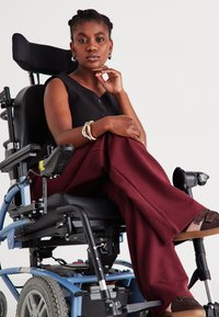 Fauteuil roulant électrique avec un cadre bleu, un siège noir et des roues grises. Le modèle porte un haut noir et un pantalon bordeaux, avec un bracelet en or.