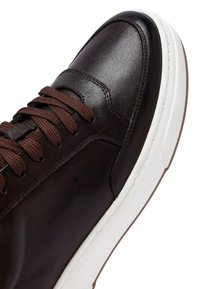Brązowe skórzane sneakersy z okrągłym noskiem, kontrastującą białą podeszwą, brązowymi sznurówkami i subtelnymi detalami szycia w górnej części.