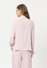 Hellrosa Bluse mit langen Ärmeln, glatter Textur, Rückenfalte und Knopfdetails an den Manschetten, kombiniert mit passenden Hosen.