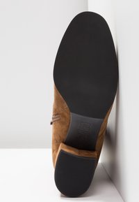 Chaussure en daim marron reposant sur le côté, montrant une semelle noire lisse et un talon carré sur fond blanc.
