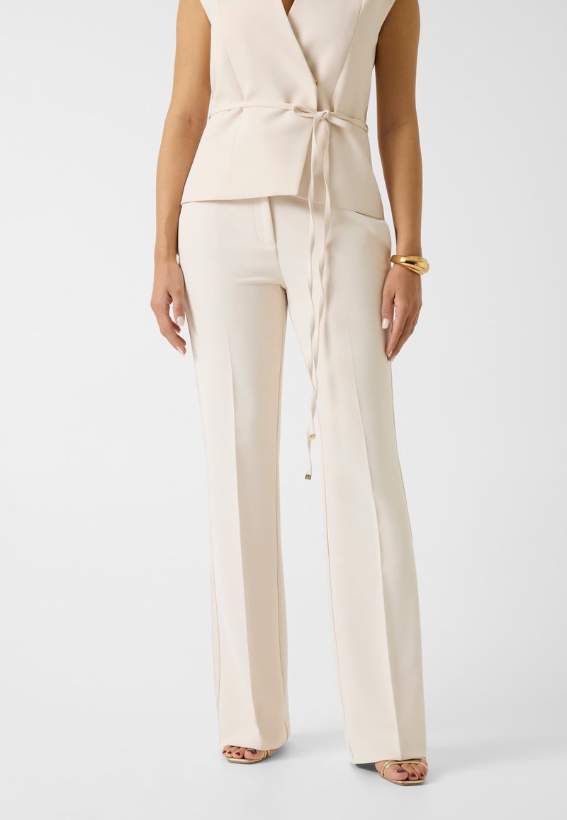 Femme portant un haut cache-cœur sans manches crème et un pantalon large assorti, avec un bracelet doré et des talons ouverts, debout sur fond blanc.