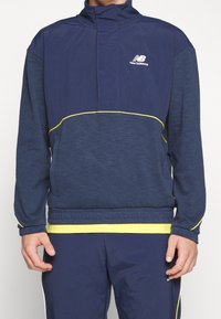 Tmavě modrá sportovní bunda s vysokým límcem, žlutými pásky jako akcentem a strukturovanou tkaninou. Na hrudi je logo značky New Balance.