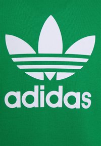 Grön bomulls t-shirt med en stor vit Adidas-logotyp som har tre ränder och bladliknande detaljer ovanför varumärket.