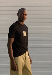 Μαύρη βαμβακερή μπλούζα με υφασμάτινο χρυσό ορθογώνιο σχέδιο στο στήθος, σε συνδυασμό με ανοιχτό μπεζ παντελόνι. Απλός λαιμός με στρογγυλή λαιμόκοψη.