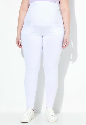 Schwangere Person trägt weiße Umstandsleggings und weiße Turnschuhe, steht vor einem schlichten hellen Hintergrund.