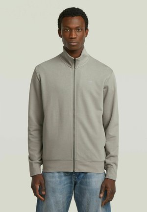 Jonge man die een grijze jas met rits en hoge kraag draagt, gecombineerd met lichtblauwe spijkerbroek, staand tegen een effen lichte achtergrond.