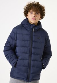 Chaqueta acolchada azul marino con capucha, que cuenta con cierre de cremallera, bolsillos laterales y un diseño acolchado para mayor textura.