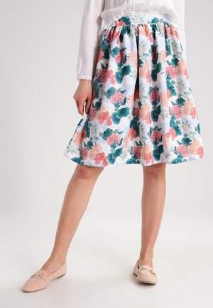 Kvinde iført et blomsterprintet nederdel til knæet med rosa, grønne og ferskenfarvede blomster, sat sammen med en hvid bluse og lys rosa flade sko.