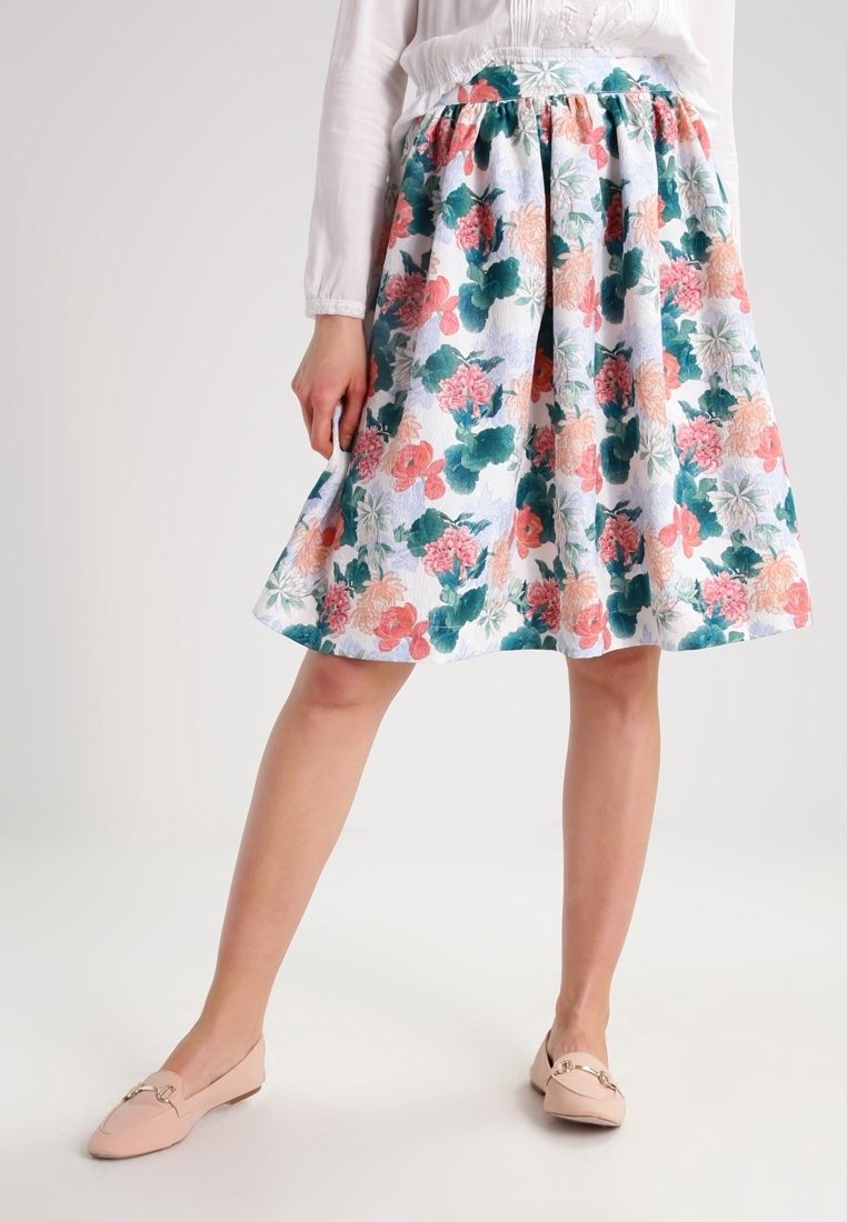 Femme portant une jupe fleurie mi-longue avec des fleurs roses, vertes et pêches, associée à un chemisier blanc et des chaussures plates rose pâle.