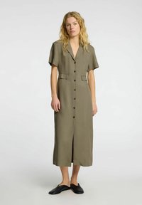 Oliven grøn midi-kjole med korte ærmer, knaplukning foran, to frontlommer og en slids i kanten. Bæret med sorte slip-on sko.