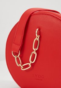 Czerwona okrągła torebka z teksturowanego materiału syntetycznego, z złotym łańcuszkowym paskiem i wytłoczonym logo na przodzie.