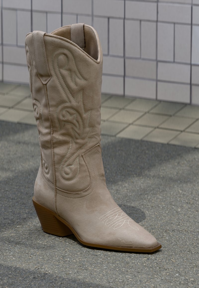 Μπεζ σουέντ καουμπόικη μποτά με μυτερή μύτη, λεπτομερή κέντημα και μέτριο στοίβαγμα τακουνιού. Λεπτή υφή και δύο λουράκια για εύκολη εφαρμογή.