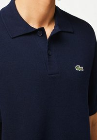 Polo bleu marine en tissu tricoté texturé, doté d'un col et d'une patte à deux boutons. Petit logo vert sur la poitrine gauche.