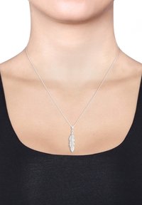 Silberner Federanhänger an einer dünnen Kette, mit detaillierter Federstruktur, getragen auf nackter Haut mit einem schwarzen Oberteil.