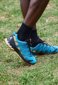 Persona che indossa scarpe da trail running blu e nere con calze nere, in piedi su erba verde all'aperto.