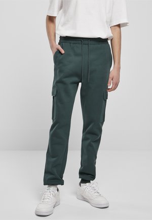 Personne portant un pantalon jogger cargo vert foncé avec poches latérales, des baskets blanches, des chaussettes blanches et une chemise blanche à manches courtes ample, debout avec une main dans la poche.
