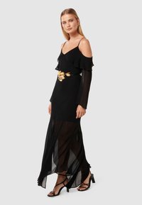 Čierne šifónové šaty s off-shoulder volánikmi, zlatý pásik v tvare kvetu a priesvitný presah; skombinované s čiernymi sandálmi na podpätku.