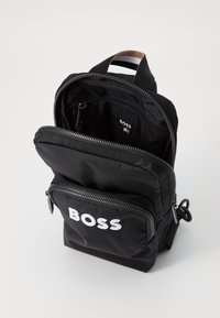 Crni najlon ruksak s više zip zatvorenih pregrada, s istaknutim bijelim „BOSS“ logotipom sprijeda i smeškim ručkom.