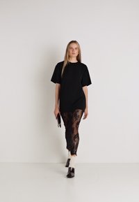 Czarna oversizeowa koszulka zestawiona z przezroczystymi czarnymi legginsami z koronkowymi wzorami kwiatowymi. Modelka nosi czarne buty i trzyma kopertówkę.