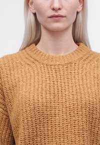 Jeune femme à la peau claire et aux taches de rousseur portant un pull en maille texturée de couleur moutarde, visible du cou à la poitrine.