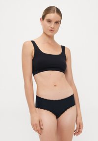 Kvinna i svart ribbat bralette och matchande högskurna trosor med vågiga kanter, stående mot en ljus, enhetlig bakgrund.