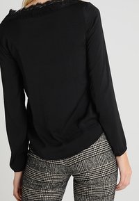 Schwarzes Langarmoberteil mit Spitzenbesatz am Ausschnitt, kombiniert mit grauen karierter Hose aus strukturierter Stoff und tailliertem Design.