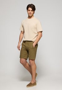 Beige t-shirt med "Polo Club" broderi, olivgröna shorts och ljusbruna espadrillos. Modellen står mot en neutral bakgrund.