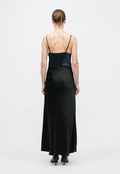 Kobieta z włosami związanymi w kok, ubrana w czarny satynowy top na cienkich ramiączkach, długą czarną spódnicę i czarne buty na obcasie, stojąca na białym tle.