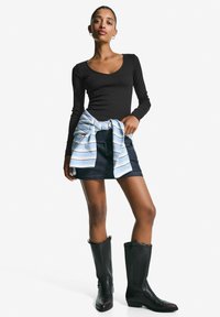 Schwarzes langärmeliges Oberteil, kombiniert mit einem dunkelblauen Jeansrock. Ein hellblau gestreiftes Hemd ist um die Taille gebunden. Schwarze kniehohe Stiefel.