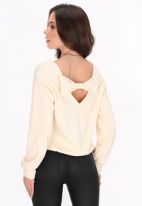 Maglia a maniche lunghe color crema con un dettaglio a fiocco sul retro e un'apertura a V. Tessuto morbido, vestibilità rilassata, abbinata a pantaloni neri attillati.