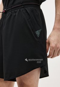 Μαύρο αθλητικό σορτς με πλαϊνή τσέπη, ελαστική μέση με κορδόνι και λογότυπο "Klättermusen Diser" κοντά στο τελείωμα, σε ένα άτομο που στέκεται.