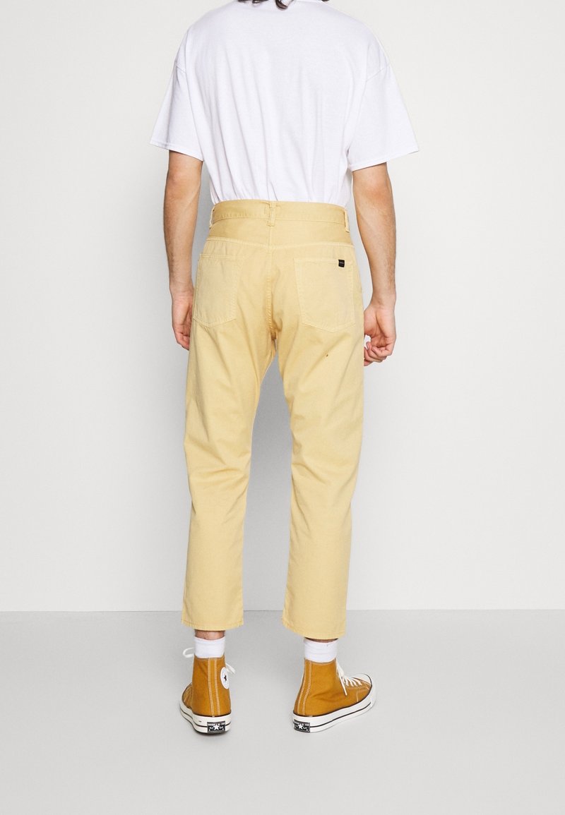 Pantalon en coton beige à coupe droite, deux poches arrière et un détail avec étiquette. Associé à des baskets montantes jaunes et des chaussettes blanches.