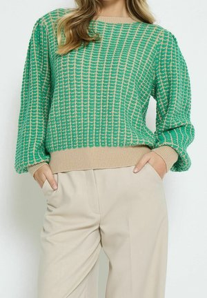 Kobieta ubrana w zielony i beżowy sweter z teksturowanym dzianinowym wzorem z balonowymi rękawami oraz beżowe spodnie z wysokim stanem, z rękami w kieszeniach.