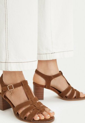 Braune Wildleder-Sandalen mit T-Riemen und Blockabsatz, getragen zu weißen, gesteppten Weitbein-Hosen vor hellem Hintergrund.