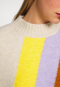 Gros plan sur une personne portant un pull crème à côtes avec de larges rayures verticales jaunes, lavande et marron au niveau du col.