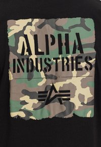 Τετράγωνο καμουφλάζ με μαύρο κείμενο "ALPHA INDUSTRIES" και λογότυπο αστέρι σε μαύρο υφασμάτινο φόντο.