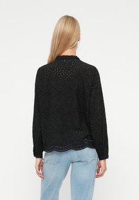 Svart brodert bluse med lange ermer, strukturert mønster, bølget kant, med øyelettdetaljer, kombinert med lyseblå jeans.