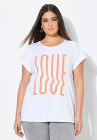 Witte korte mouwen t-shirt met oversized oranje "LOVE" tekstontwerp. Zachte stof, ronde halslijn, losse pasvorm. Gecombineerd met grijze jeans.