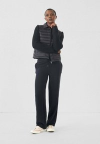 Zwarte puffer gilet over een zwarte sweater, gecombineerd met losse zwarte broek en lichtgekleurde sneakers. Glad textuur met een slanke silhoutte.