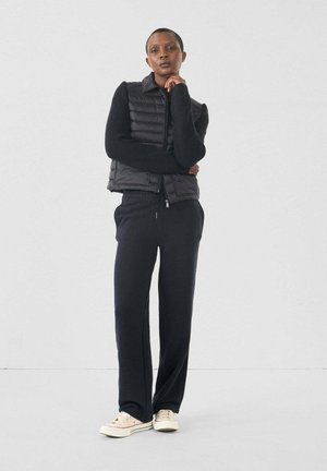 Zwarte puffer gilet over een zwarte sweater, gecombineerd met losse zwarte broek en lichtgekleurde sneakers. Glad textuur met een slanke silhoutte.