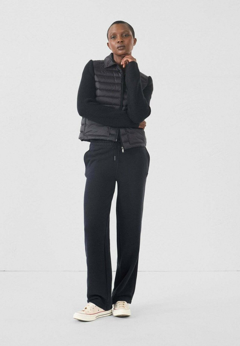 Chaleco acolchado negro sobre un suéter negro, combinado con pantalones negros holgados y zapatillas de color claro. Texturas suaves con una silueta elegante.