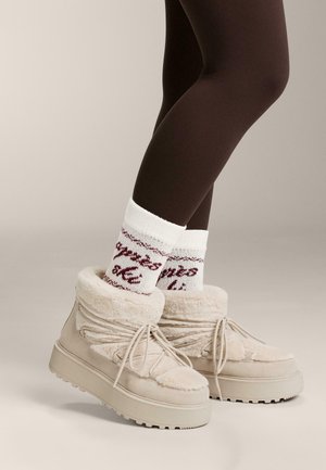 Cizme bej căptușite cu blană, cu tălpi groase, cu șireturi și textură moale. Accesorizate cu șosete albe care afișează un text cu model roșu.