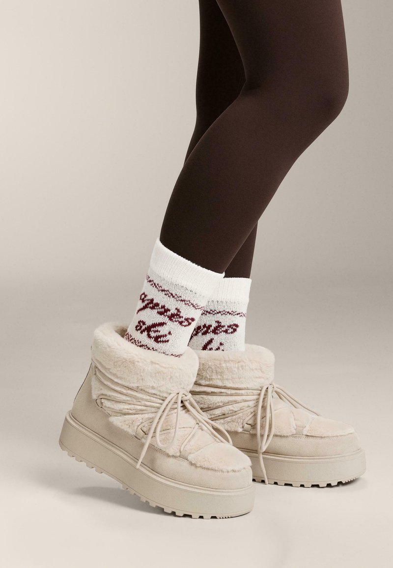 Beżowe kozaki wyłożone futerkiem, z masywną podeszwą, sznurowane i o miękkiej fakturze. Uzupełnione białymi skarpetkami z czerwonym, wzorzystym napisem.