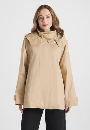 Vrouw draagt een beige capuchonjas met hoge kraag en knoopdetails, gecombineerd met zwarte broek tegen een effen witte achtergrond.