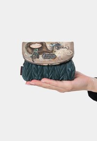 Bolso texturizado verde con una solapa estampada que presenta una ilustración y la palabra "Sueños". Diseño compacto sostenido en una mano.