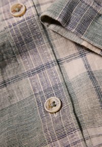 Camisa de cuadros de algodón con líneas verdes y azules sobre un fondo beige, con cuello redondeado y un detalle de botón crema.